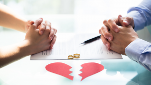 Hands of husband and wife on table next to broken heart, wedding rings, and divorce decree paper.